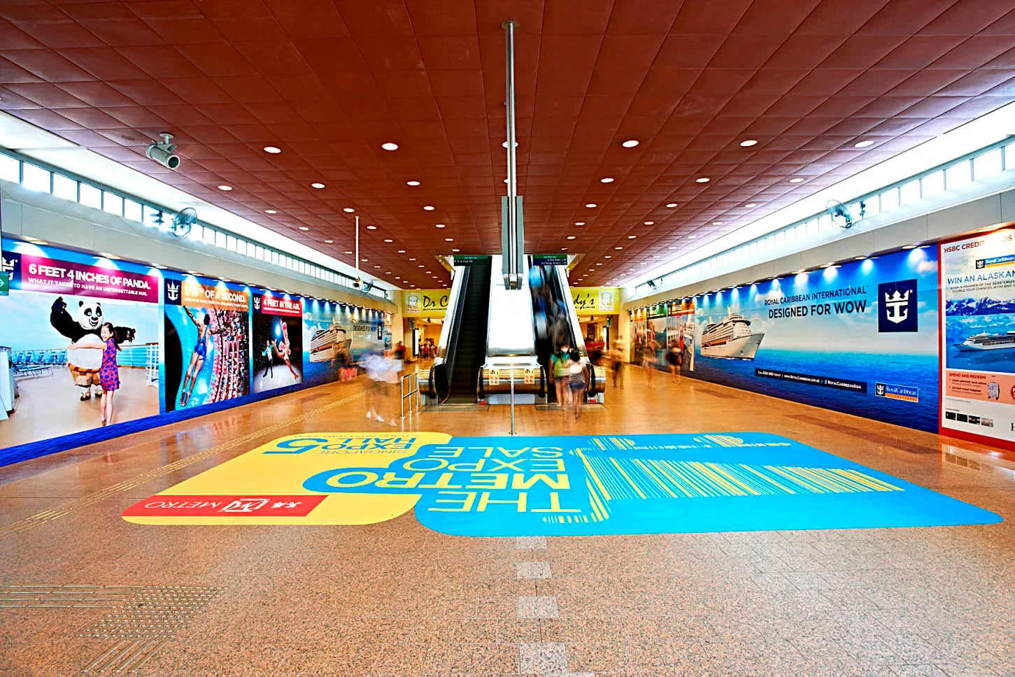 Indoor shopping mall corridor with escalators, colorful floor graphics, and advertising banners