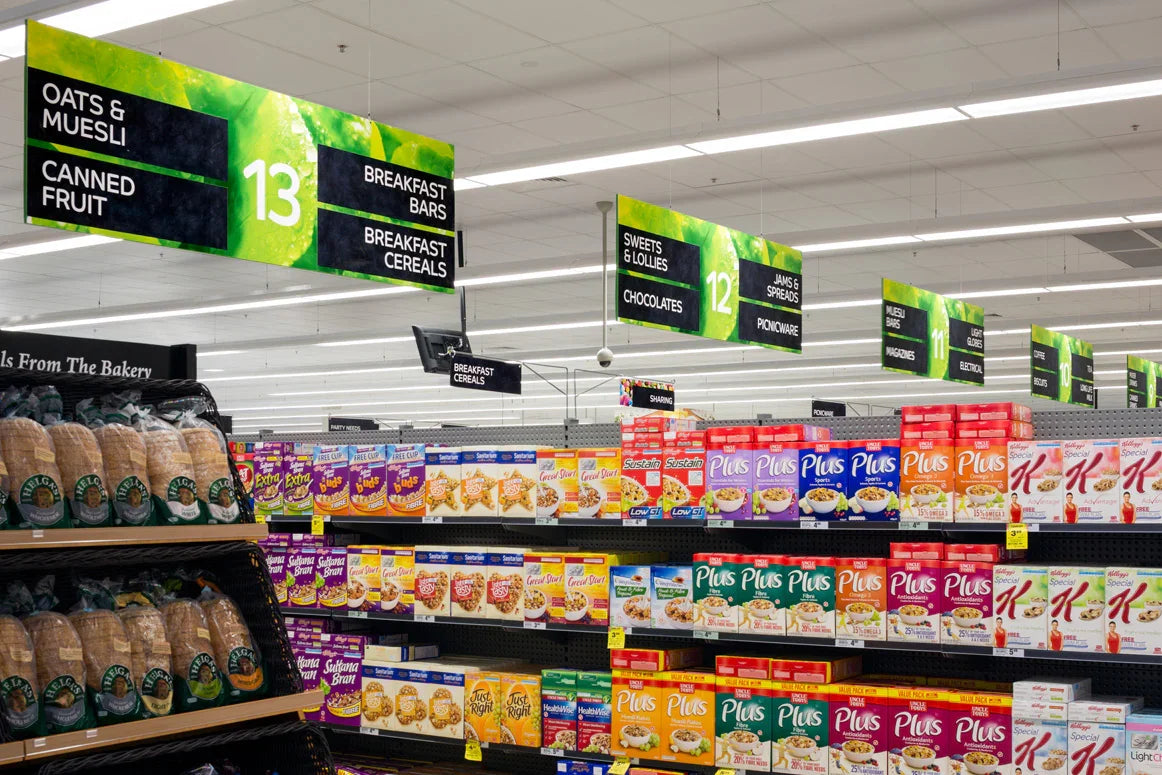 Supermarket aisle with bread, breakfast cereal boxes, and aisle signs for oats, muesli, sweets.